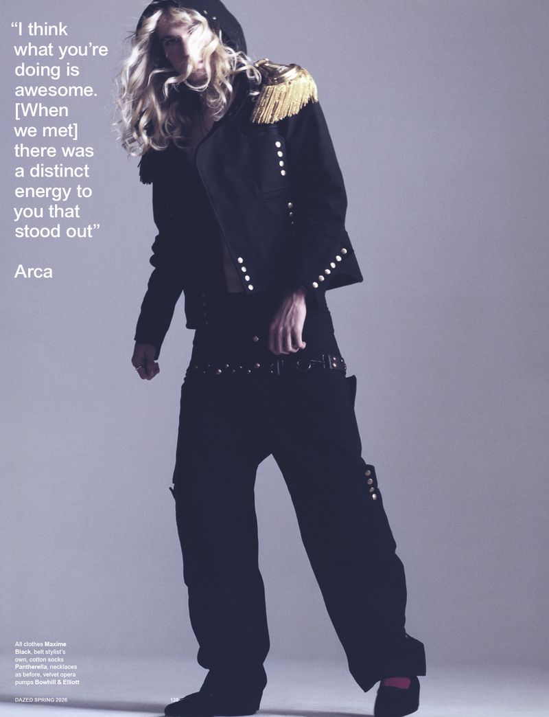 Model poses in a black military-style jacket with gold epaulettes, loose black pants, and black shoes against a neutral background.