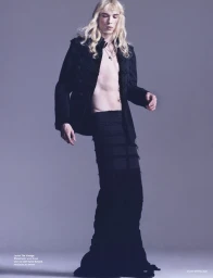Model with long blonde hair wearing a black jacket and long black skirt against a grey studio background