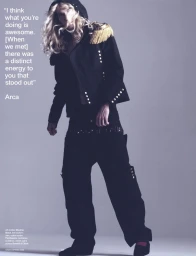 Long-haired blonde person wearing a black military-style jacket with gold epaulettes and dark trousers against a grey backdrop.