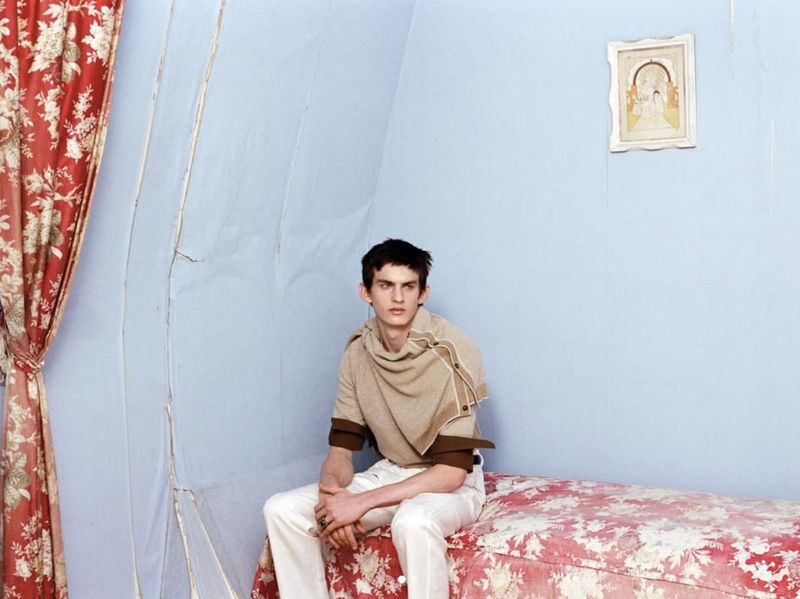 Young man sits on a bed in a blue room with red floral curtains. He wears a tan sweater, white pants, and a ring. Framed art hangs on the wall.