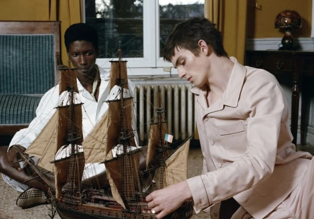 Two people, an adult male and adult female, sit in an interior with a model sailboat, wearing matching pajamas, in soft indoor lighting.