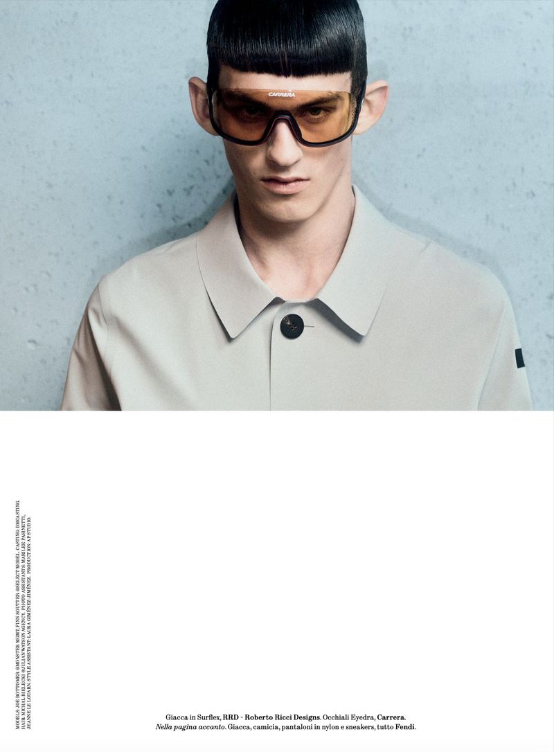 Portrait of a male model wearing tinted wraparound sunglasses and a light grey collared shirt against a mottled blue-grey background.