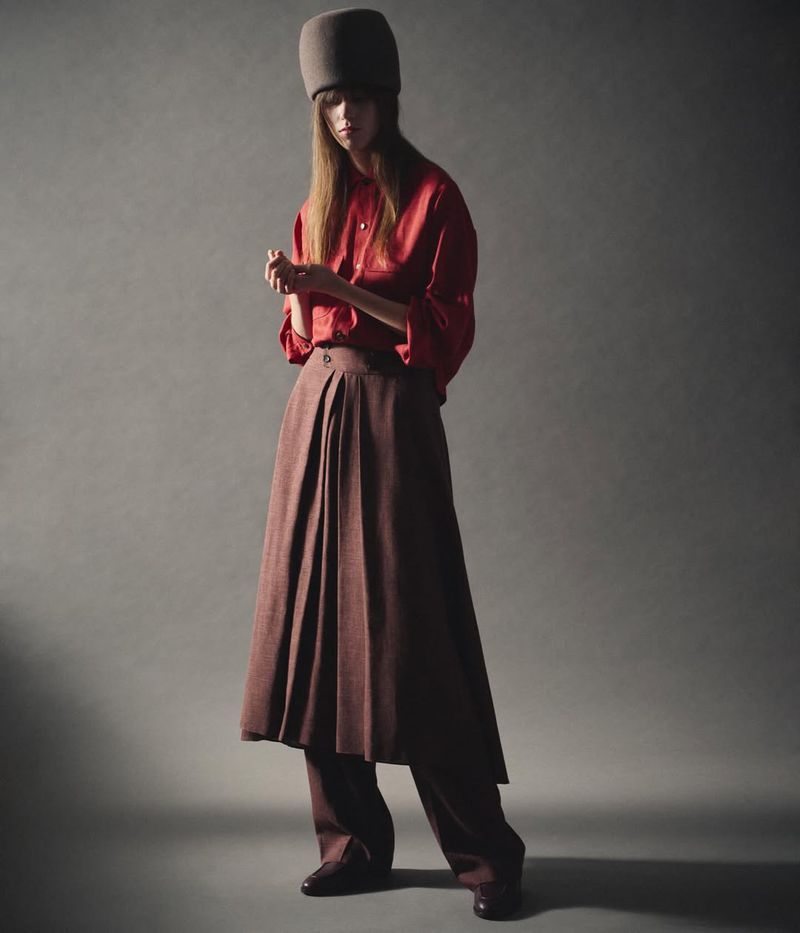Woman in a red silk shirt, brown skirt with pants underneath, and a brown hat poses in a studio against a neutral gray background.