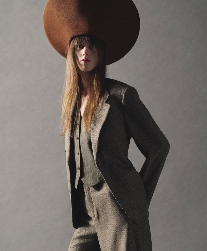 Model wearing a wide-brimmed brown hat and grey suit jacket against a neutral grey studio background.
