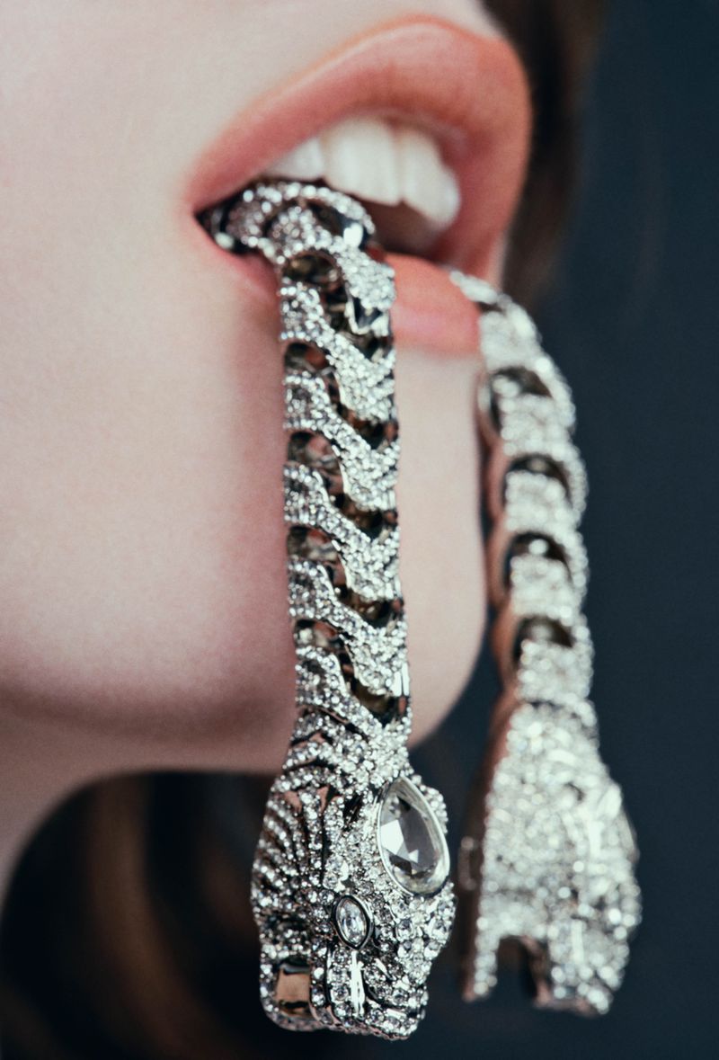 Close-up of a person with pink lips holding sparkling silver snake-shaped jewelry in their mouth against a dark grey background.