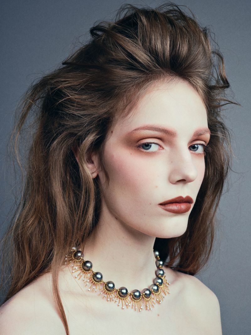 Close-up shot of a young adult female model with fair skin and brown hair, wearing a pearl necklace, against a grey background.