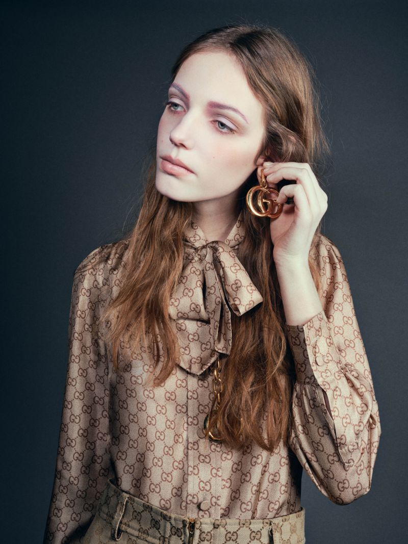 A fashionable young woman with long brown hair wears a Gucci blouse and earrings against a muted gray backdrop in a studio portrait.