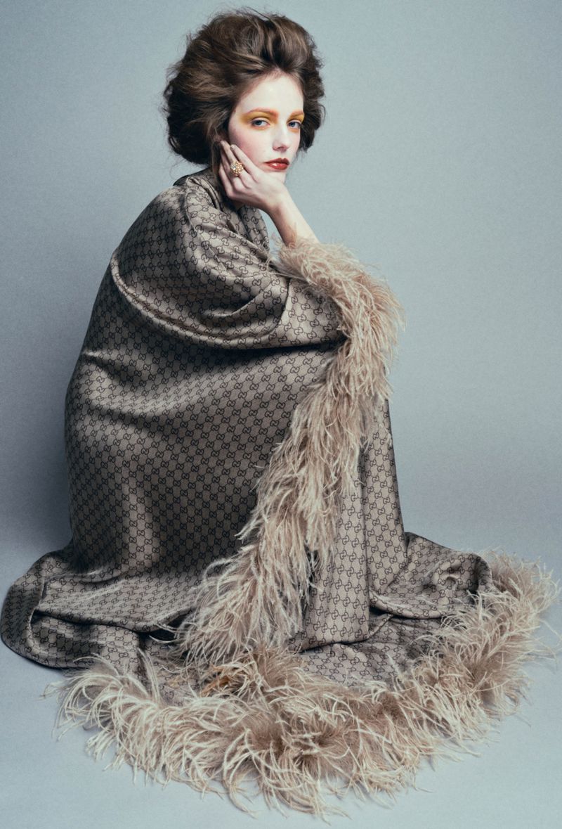 Woman in patterned coat with feather trim posing in a studio with soft, diffused lighting and neutral background for a fashion editorial.