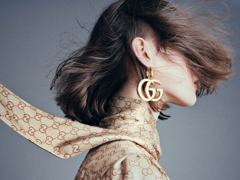 Fashion shot of a woman with wavy hair wearing a patterned top and oversized earrings with the Gucci logo.