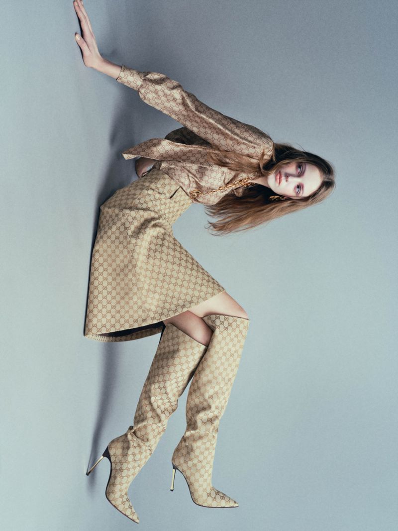Model wearing patterned beige blouse, skirt, and knee-high boots against a grey studio seamless backdrop.