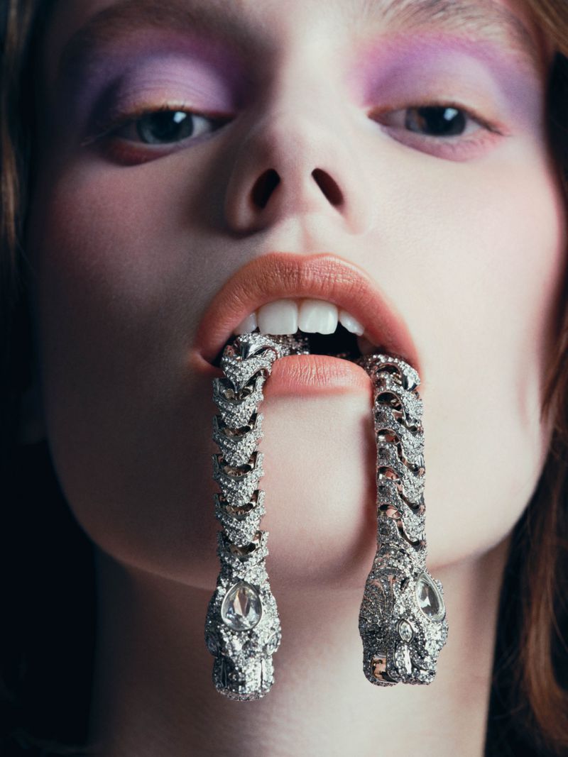 Close-up of an adult woman with purple eyeshadow and a decorative metal snake in her mouth in a studio setting.