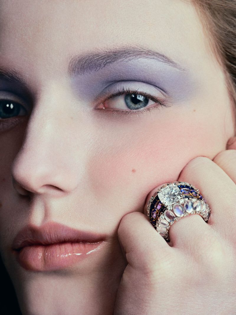 Close-up portrait of a woman wearing lavender eyeshadow and a large ring with multiple stones resting her face on her hand.