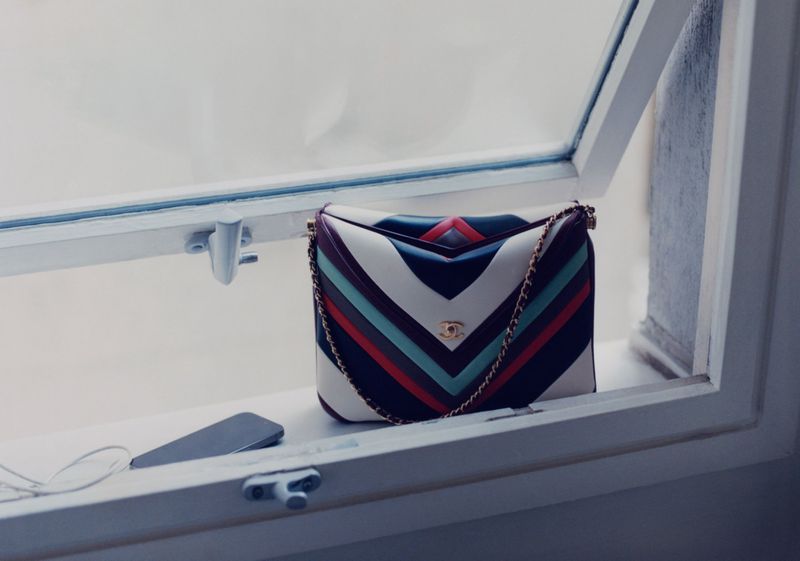 Striped Chanel handbag with gold chain, sitting on a window sill near a phone and earbuds, creating a high-fashion product shot.