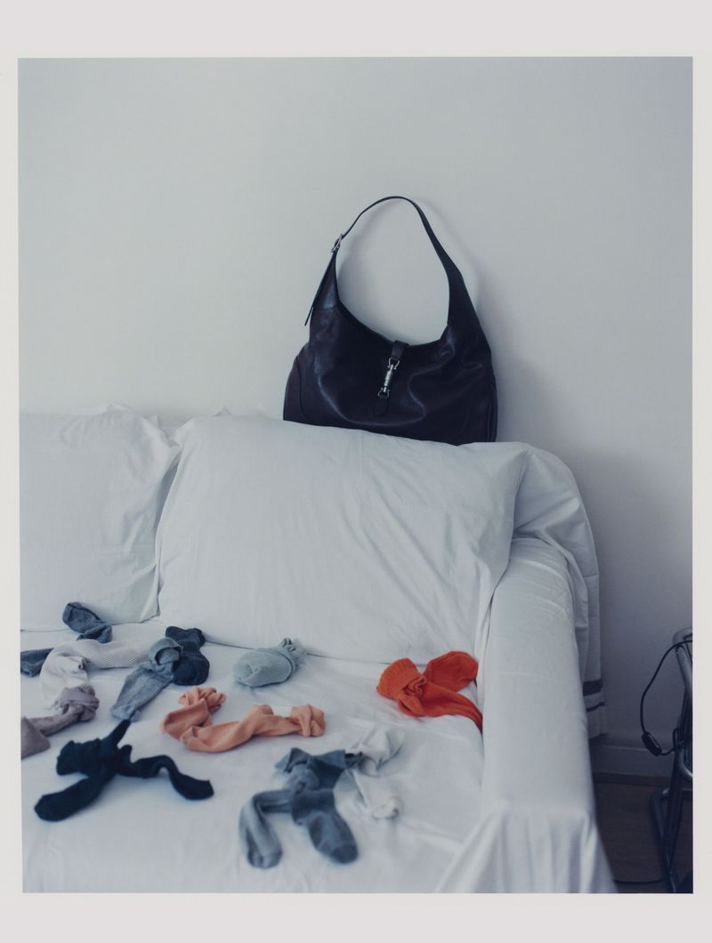Dark purse sits atop a white sofa; scattered socks create a playful foreground element against a neutral background, suggesting comfort and chaos.