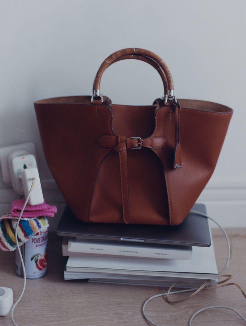 Brown leather tote bag with bamboo handles sits on books and a laptop with electrical cord plugged into a wall outlet and a soda can.