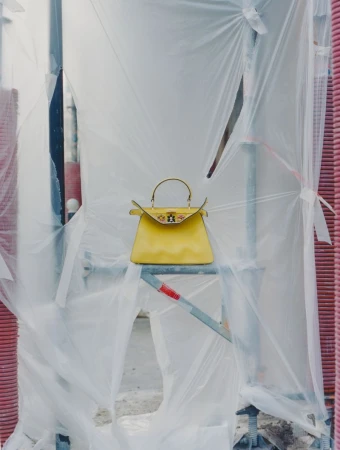 A yellow handbag is displayed on metal scaffolding covered with translucent plastic sheeting, creating a unique fashion editorial image.