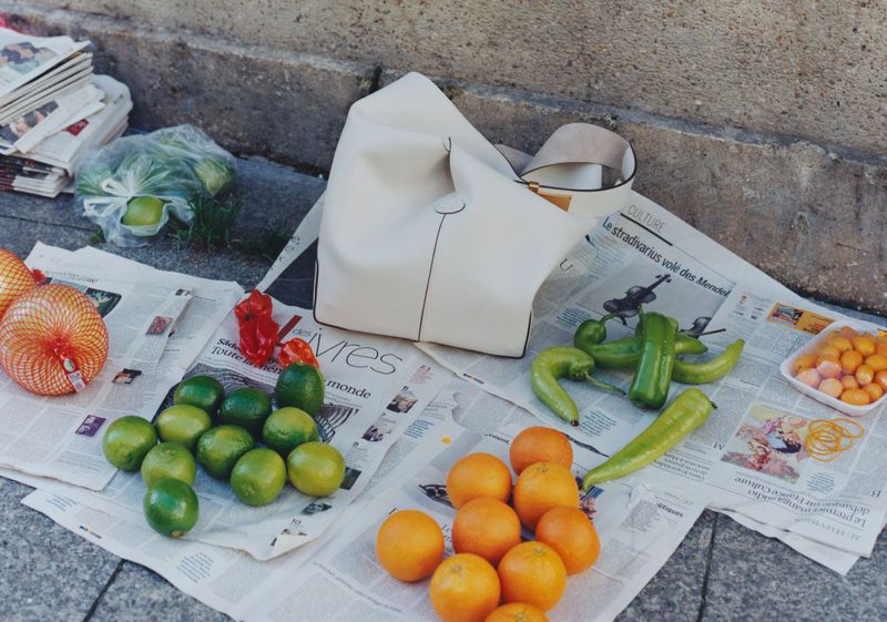 A collection of limes, oranges, peppers, and a cream-colored bag arranged on newspaper atop a concrete surface, bathed in soft, natural light.
