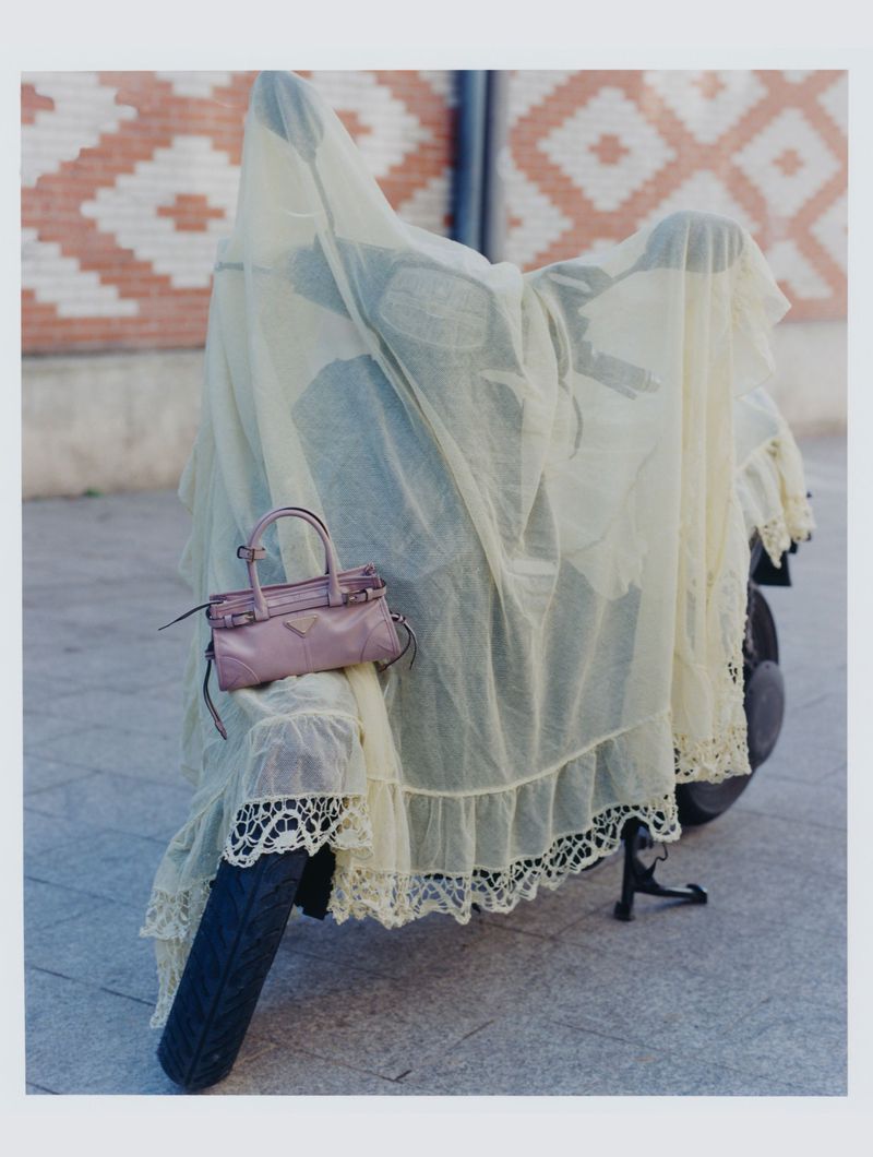 Motorcycle draped in sheer fabric with lace trim, parked on a city street with a pink leather handbag, brick-patterned wall in the background.