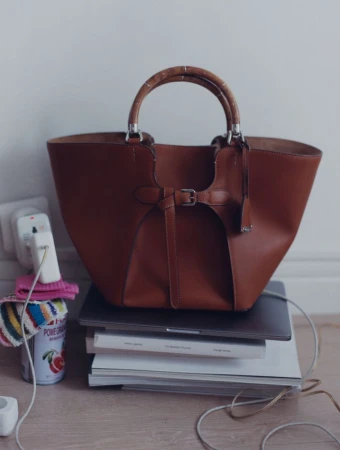 Brown leather tote bag with bamboo top handles resting on a stack of books and a laptop against a light wall.