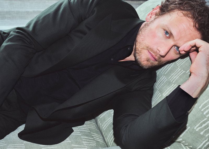Portrait of a man in a suit reclining on a patterned sofa, gazing towards the camera with a thoughtful expression in a close-up shot.
