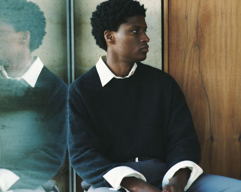An African-American man in a dark sweater with white collar, looking away. The setting includes a light-colored wood wall and mirrored surface.