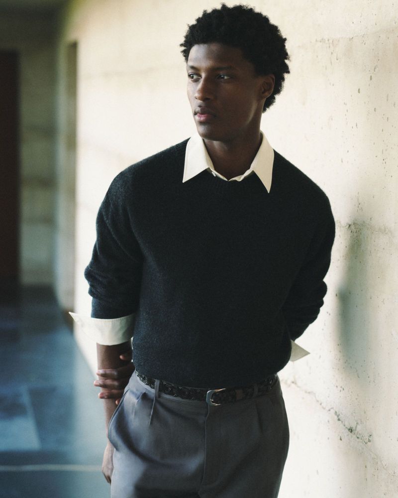 Portrait of a stylish adult Black man with dark curly hair, wearing a black sweater and grey slacks, posing indoors against a textured wall.