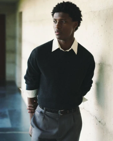 Portrait of a stylish adult Black man with dark curly hair, wearing a black sweater and grey slacks, posing indoors against a textured wall.