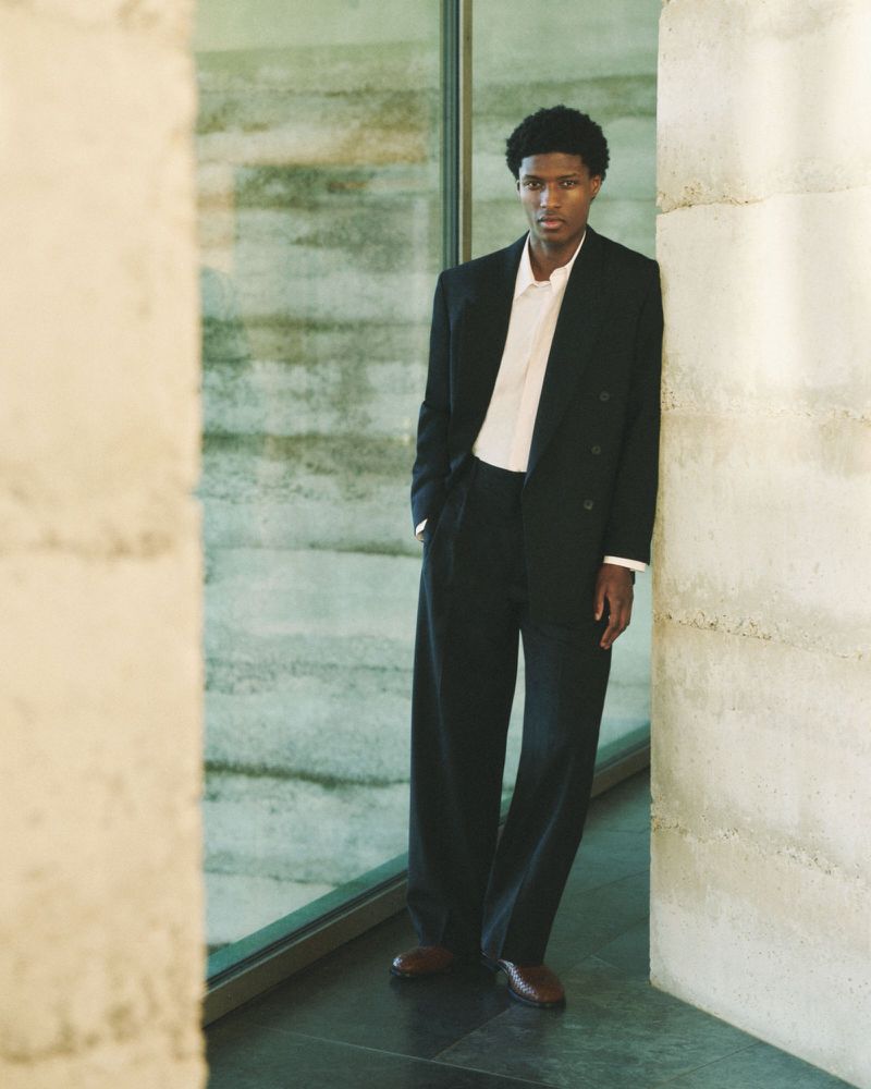 A young man in a black suit and white shirt leans against a concrete wall in a modern indoor setting, looking directly at the camera.