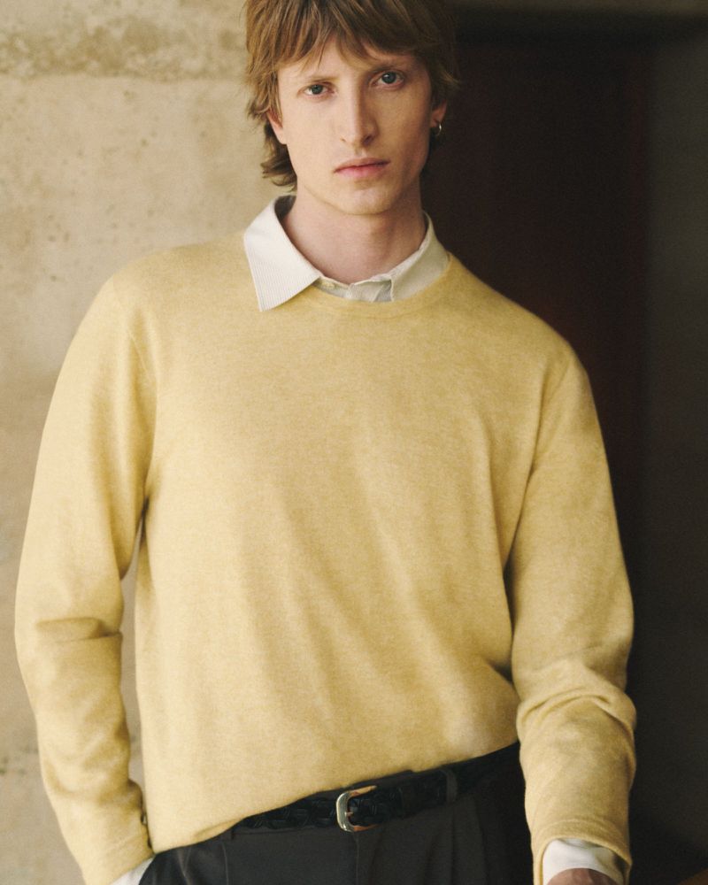 Indoor portrait of a young adult male with brown hair wearing a light yellow sweater and black pants with a neutral expression.