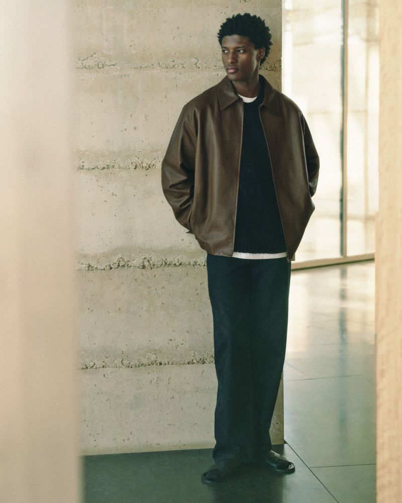 A stylish man wearing a brown leather jacket and dark blue pants poses against a textured wall in a soft, natural light setting.
