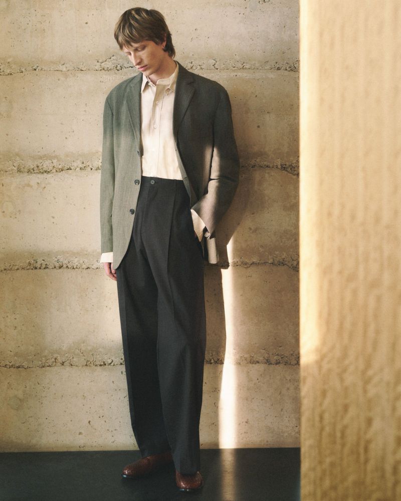 Adult man in blazer and wide-leg pants standing in front of a concrete wall. Fashion editorial style, neutral tones and soft lighting.