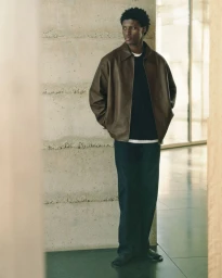 Model wearing a brown leather jacket and dark trousers standing against a textured concrete wall