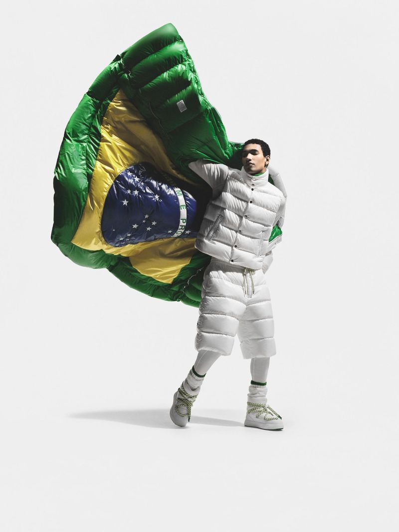Man poses in white puffer vest and shorts, holding large, inflatable Brazilian flag against a solid white backdrop in a studio shot.