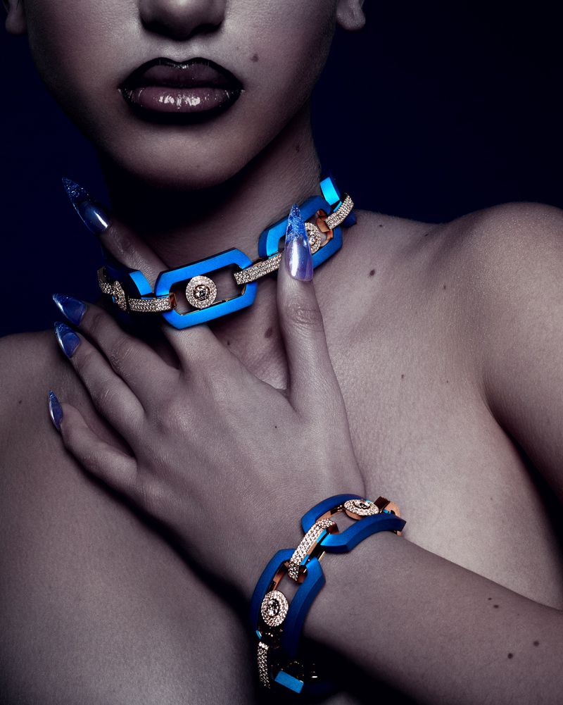 Close-up of a woman's upper torso wearing a blue link chain necklace and matching bracelet with gemstones and long blue nails.