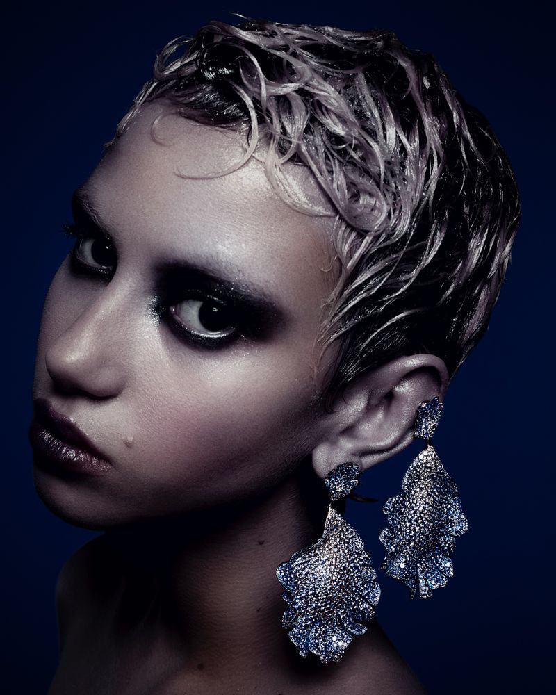 Close-up portrait of a stylish young adult female model with platinum hair, dark eye makeup, and large decorative chandelier earrings.