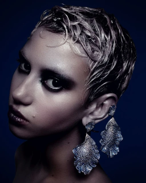 Close-up portrait of a stylish young adult female model with platinum hair, dark eye makeup, and large decorative chandelier earrings.