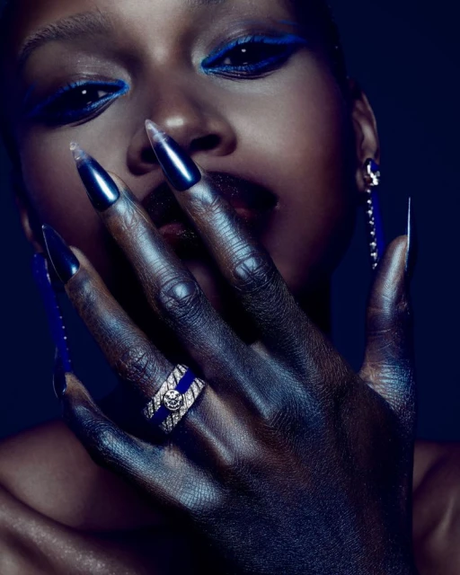 Close-up fashion shot of a Black female model with dramatic blue eye makeup, glossy dark lipstick, and long, dark, glossy fingernails.