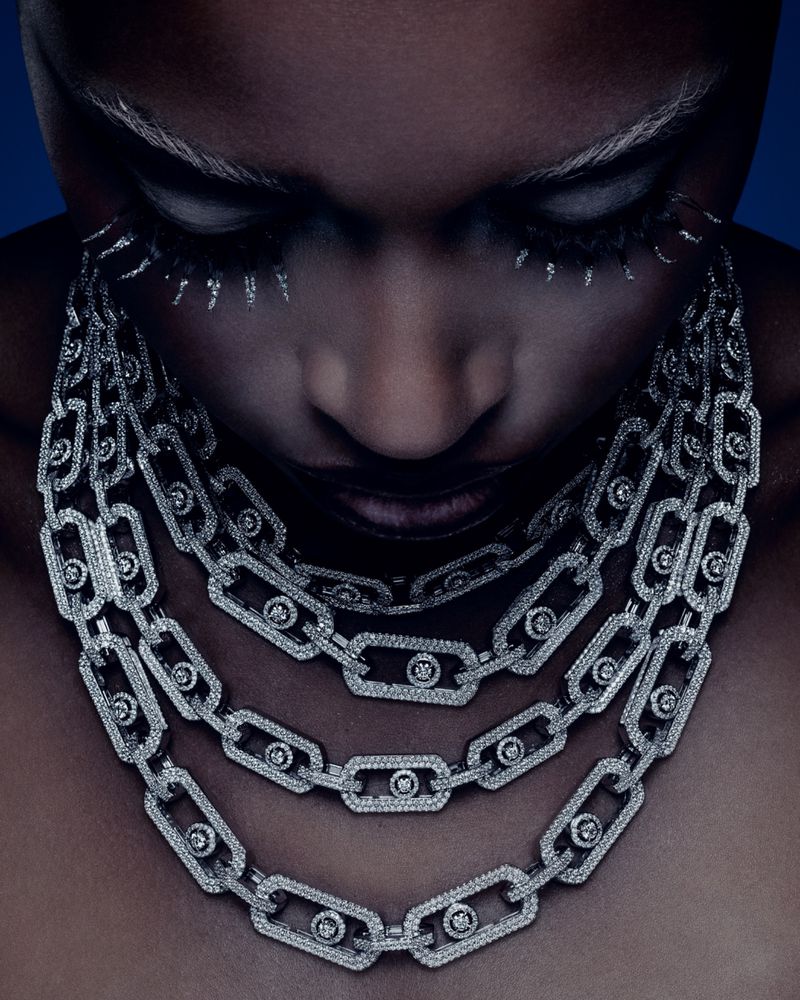 Close-up beauty shot of a Black female model with white brow and lash makeup, wearing a three-strand diamond chain necklace against blue.