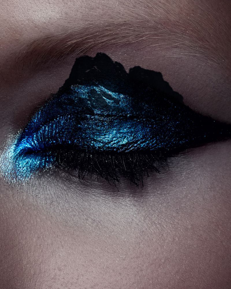 Close-up of a human eye with artistic makeup. Dark blue and black eyeshadow is applied in a wing shape with a shimmery finish.