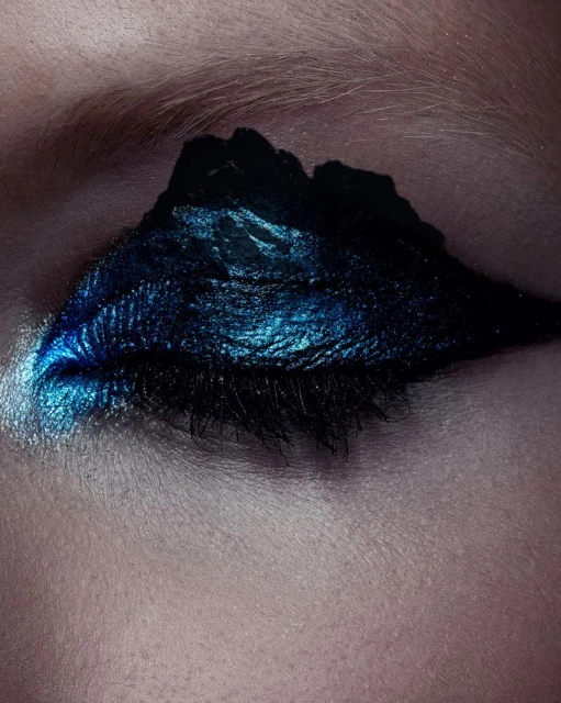 Close-up of a human eye with artistic makeup. Dark blue and black eyeshadow is applied in a wing shape with a shimmery finish.