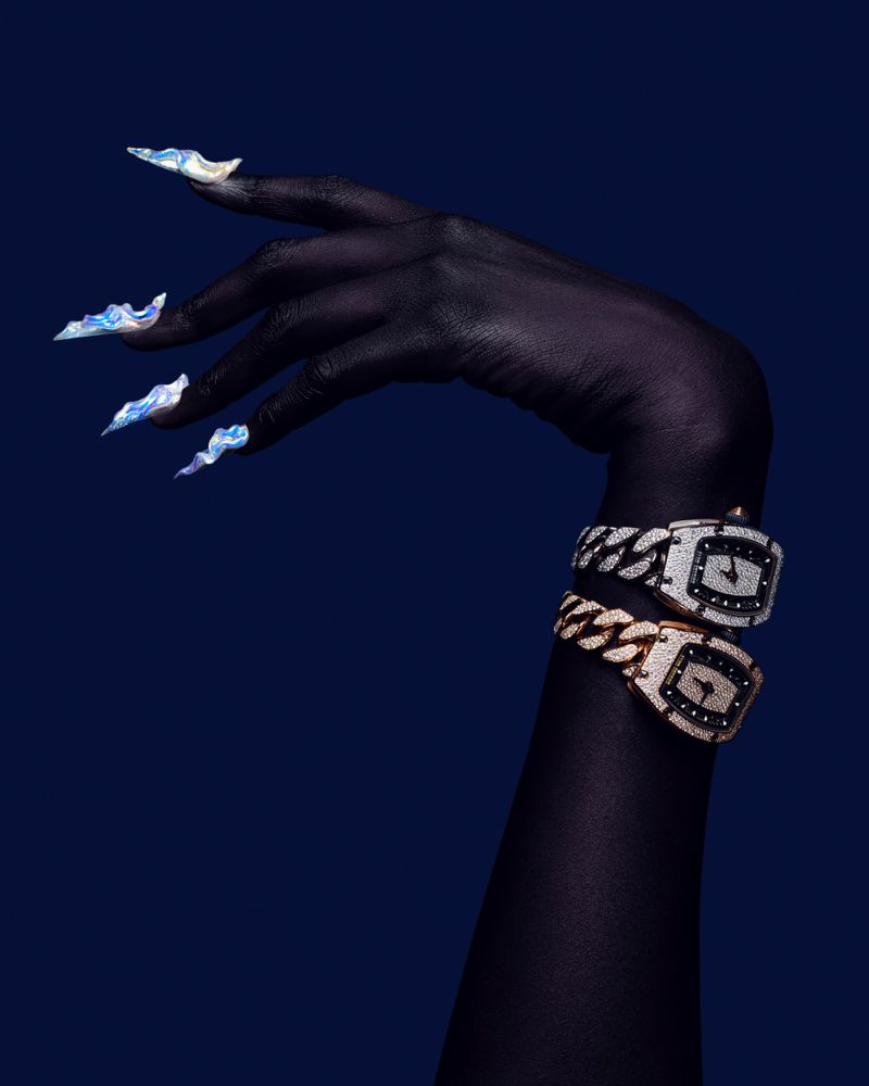 Elegant close-up of dark-skinned hand adorned with two wristwatches and statement fingernails against a deep blue backdrop.