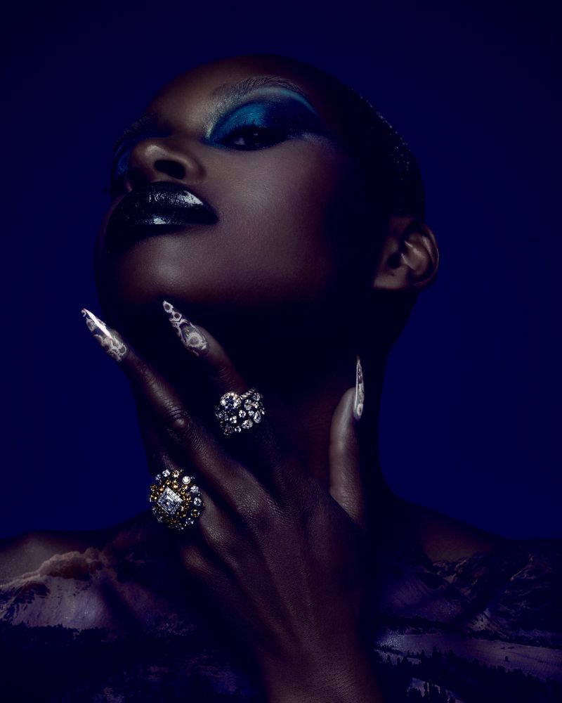 Striking beauty portrait of a Black female model with bold makeup, nail art, and statement rings against a dark blue background.