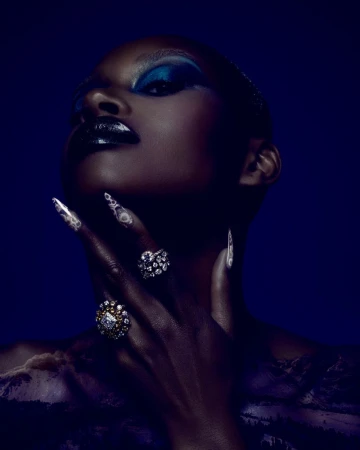 Striking beauty portrait of a Black female model with bold makeup, nail art, and statement rings against a dark blue background.