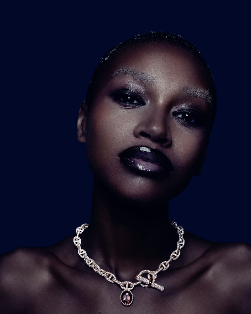 Close-up beauty shot of a woman with dark skin wearing a jeweled chainlink necklace with a pink gemstone against a navy background.