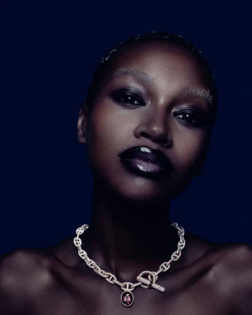 Close-up beauty shot of a woman with dark skin wearing a jeweled chainlink necklace with a pink gemstone against a navy background.