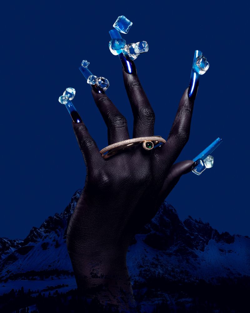 Close-up of dark-skinned hand with long blue nails decorated with ice cubes, adorned with jeweled bracelet, against blue mountain backdrop.