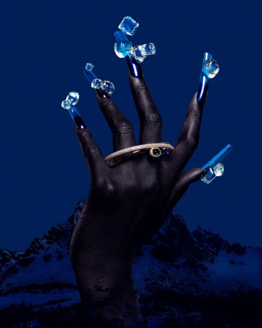 Close-up of dark-skinned hand with long blue nails decorated with ice cubes, adorned with jeweled bracelet, against blue mountain backdrop.