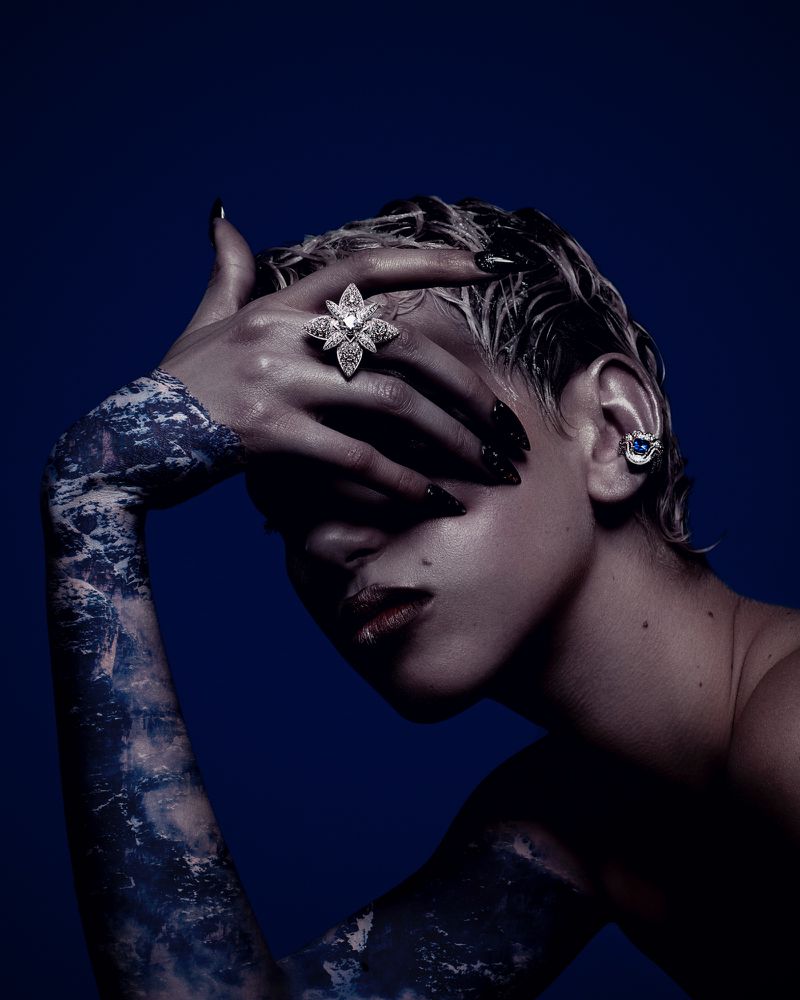 Portrait of adult woman with dramatic makeup, jewelry, and blue body paint posing against dark blue background in a beauty editorial.