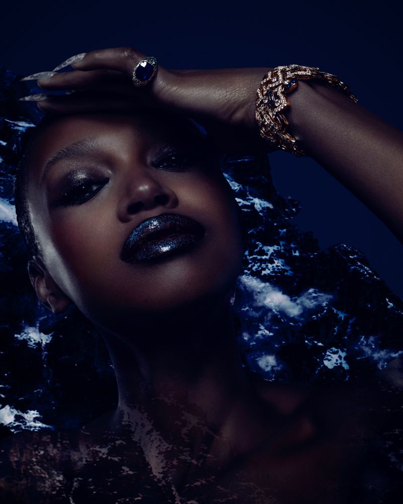 Close-up studio portrait of a woman with glitter makeup and jewelry, against a blue and white patterned backdrop. Beauty fashion.