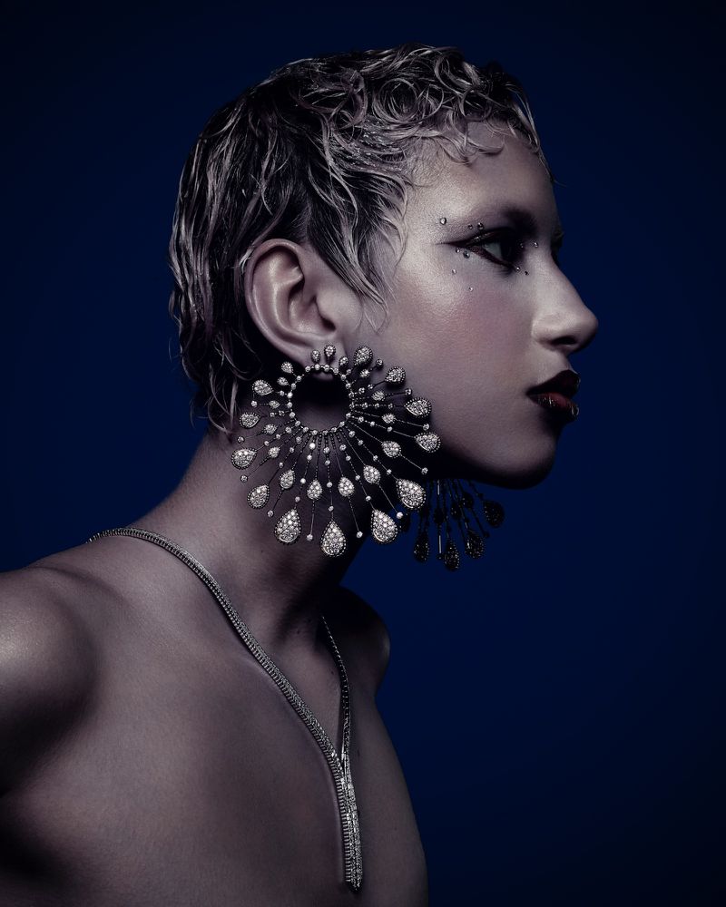 Profile shot of a woman with dramatic eye makeup and large, decorative earrings against a dark blue background in a beauty editorial.
